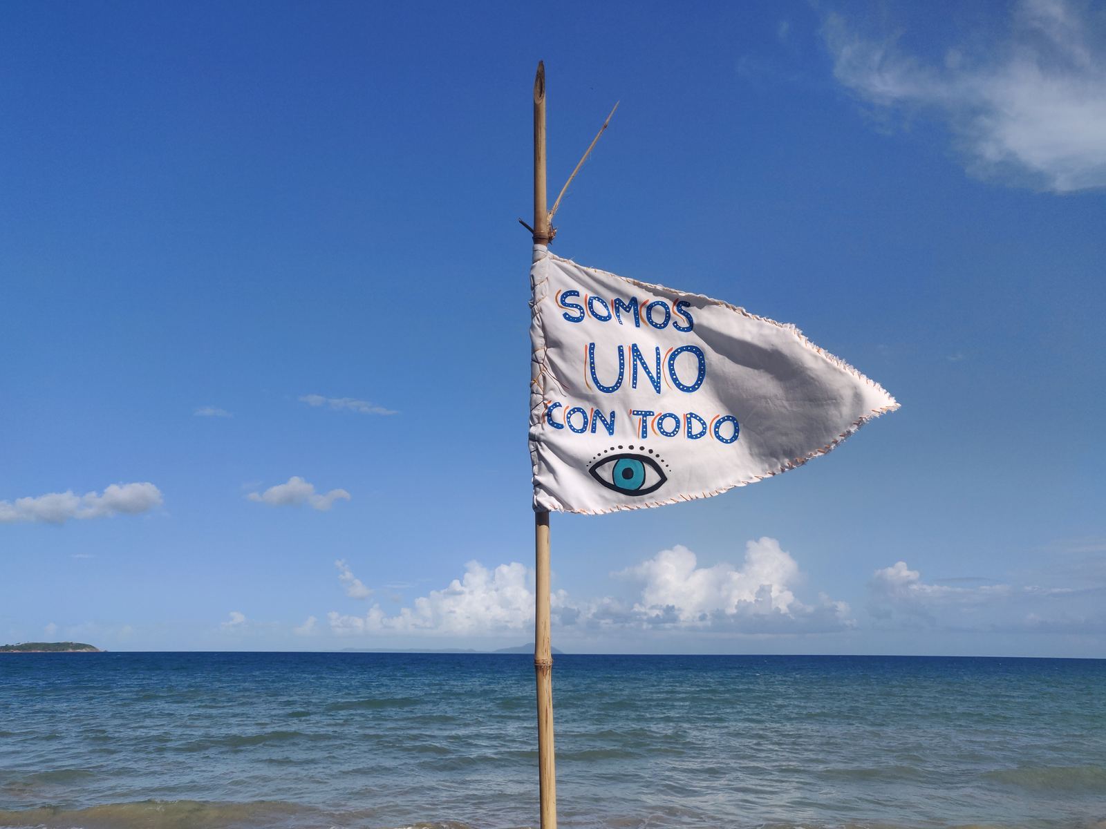 Hand-painted flag reading Somos Uno Con Todo against a bright blue ocean sky